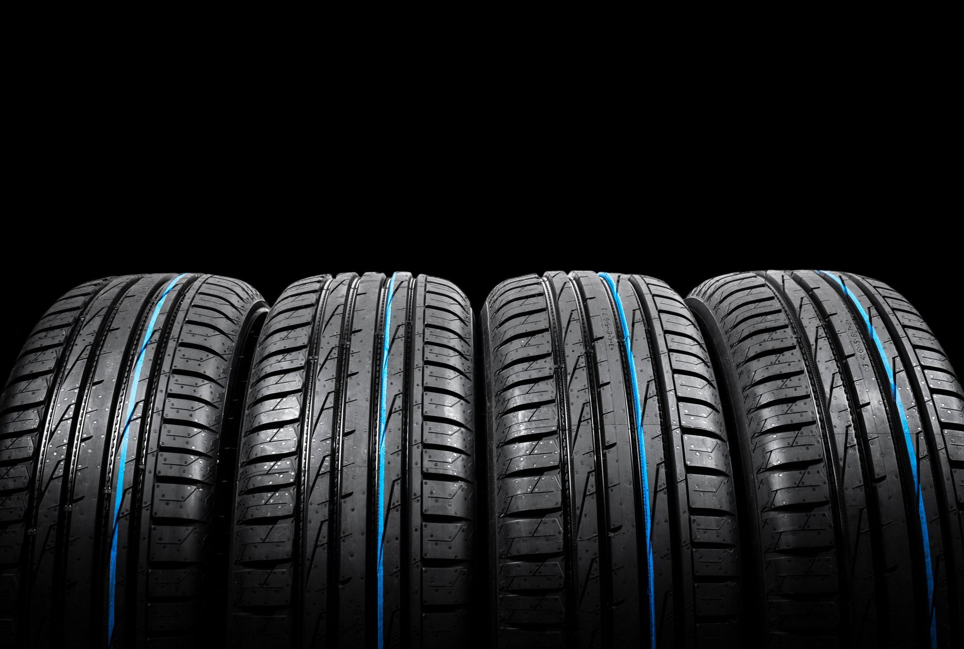 Four black rubber car tires standing side by side against a black background, each featuring a blue stripe on the tread.