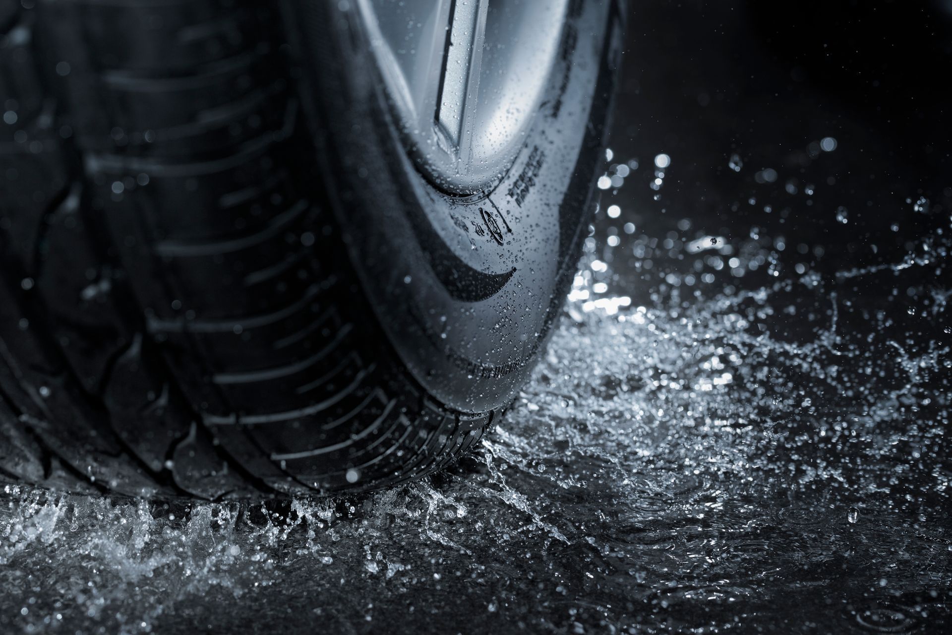 A car tire splashes through a puddle on a dark, wet road, highlighting the tread and water spray.