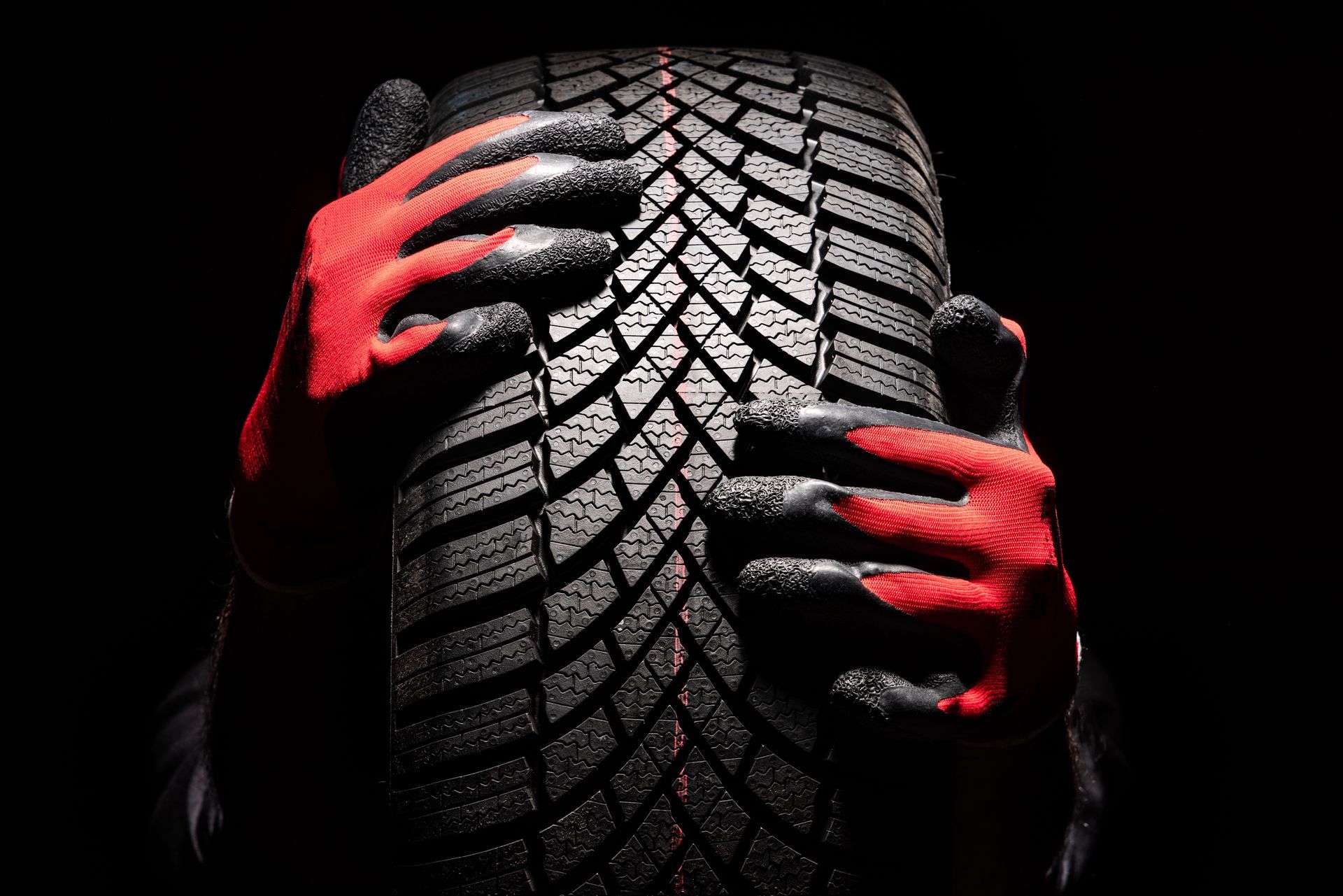 Hands wearing black and red work gloves hold a winter tire with a directional tread pattern against a black background.