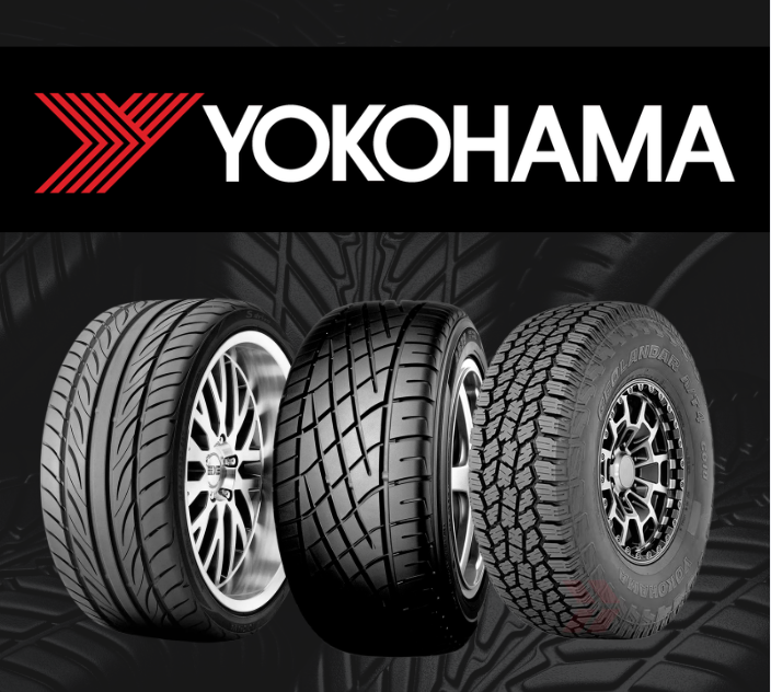 A close-up, front-facing view of a black tire with a symmetrical, aggressive tread pattern against a dark background.
