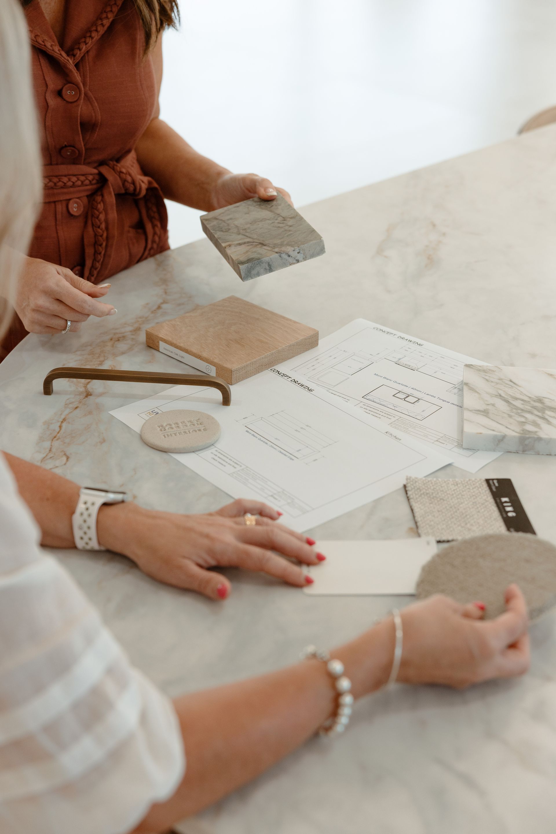 Two people at a table reviewing design samples. One holds a stone, the other holds a card. Marble table, sketches present.