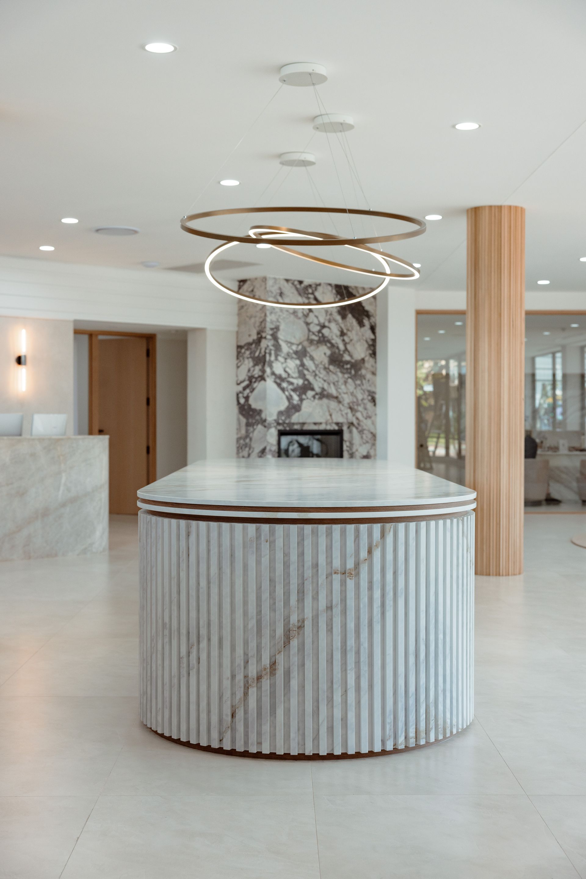 Modern, light-filled lobby with marble reception desk and fireplace. Circular light fixture, wood pillar, and white walls.