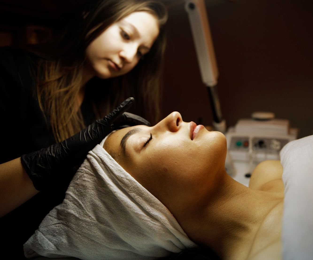 a gloved woman inspects another woman's eyebrow