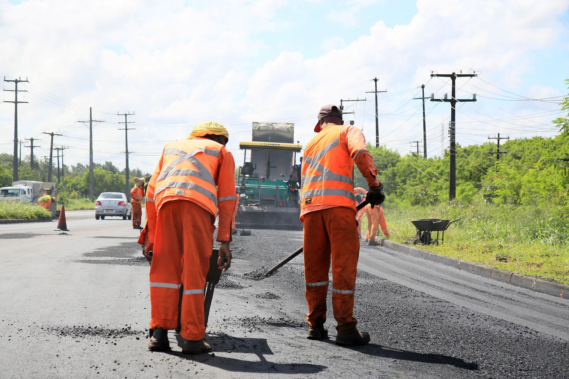 Road workers in orange vests and jumpsuits, paving a road on a sunny day.