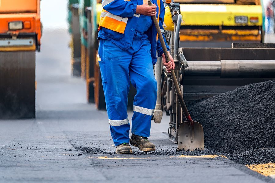 Construction worker in blue jumpsuit shoveling asphalt on a road; heavy machinery in background.