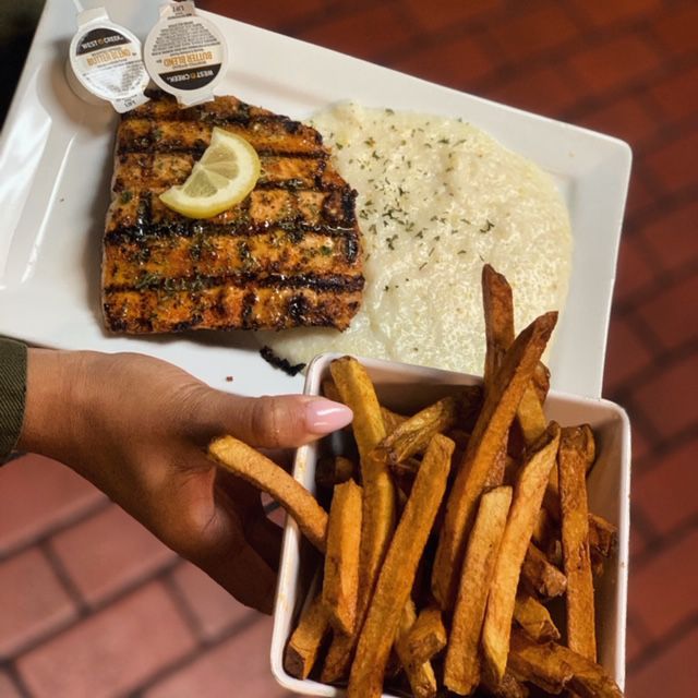 Person holding a plate with grilled salmon, grits, and sweet potato fries. 
