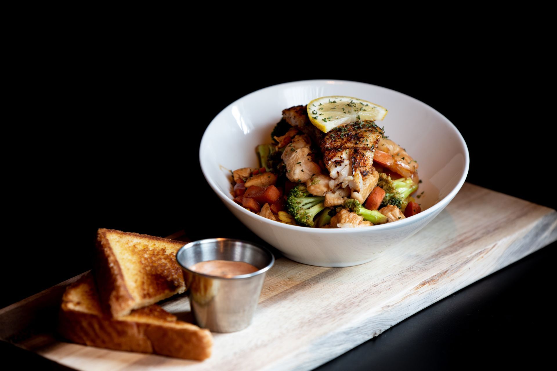 Bowl of chicken and vegetables with a lemon slice garnish, served on a wooden board with toast and dipping sauce.