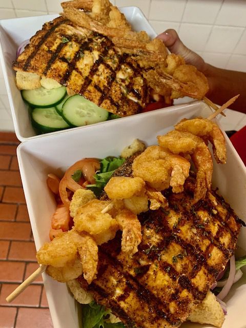 Two white bowls holding salads with grilled fish, fried shrimp skewers, and vegetables. A person holds one bowl.