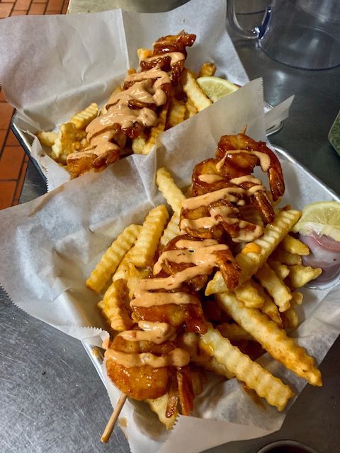 Two baskets of fries topped with shrimp skewers drizzled with sauce, served with lemon slices.