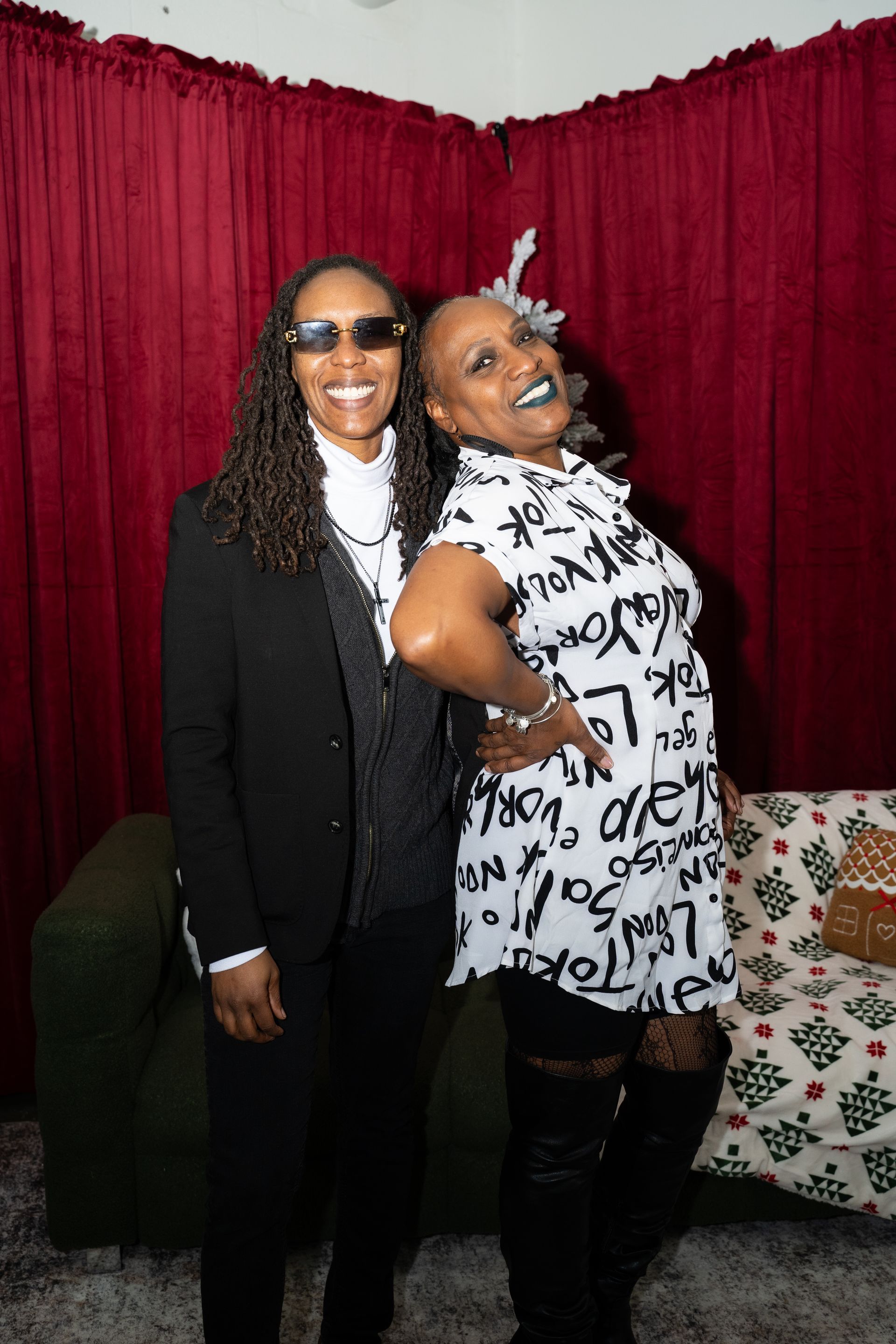 Two women pose in front of a burgundy curtain. One in a suit, the other in a white graphic dress and tall boots.