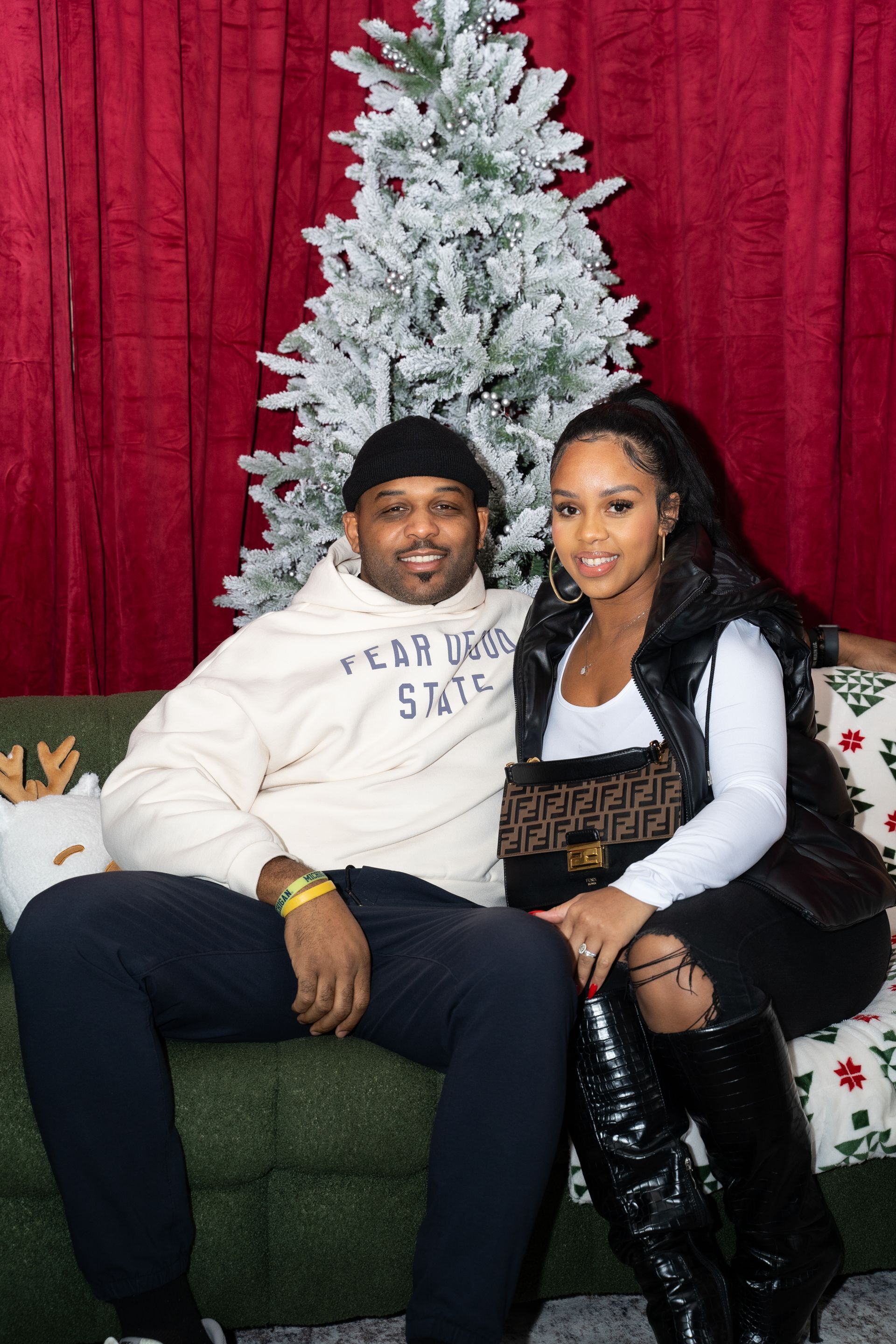 Couple sitting on a decorated couch in front of a flocked Christmas tree and red backdrop.