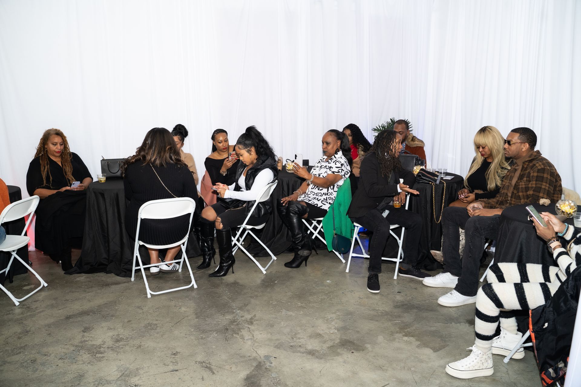 A group of people seated indoors, engaged in conversation, with a white backdrop and chairs.