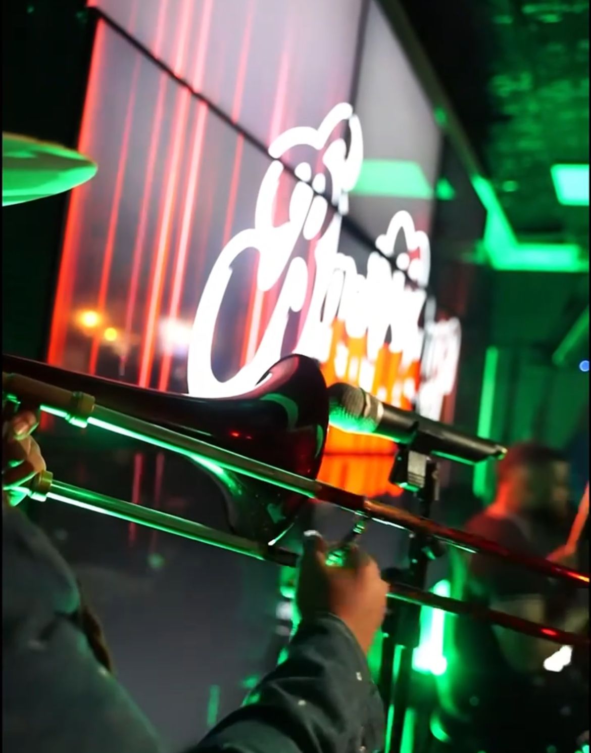 A trombone player performing in a dimly lit venue with green and red lighting. A band logo is visible in the background.