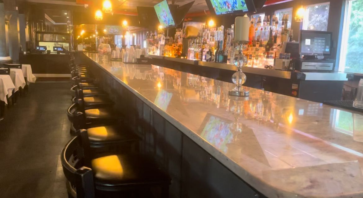 A long, marble-topped bar in a restaurant, with stools, bottles, and taps, and a glimpse of the dining area.