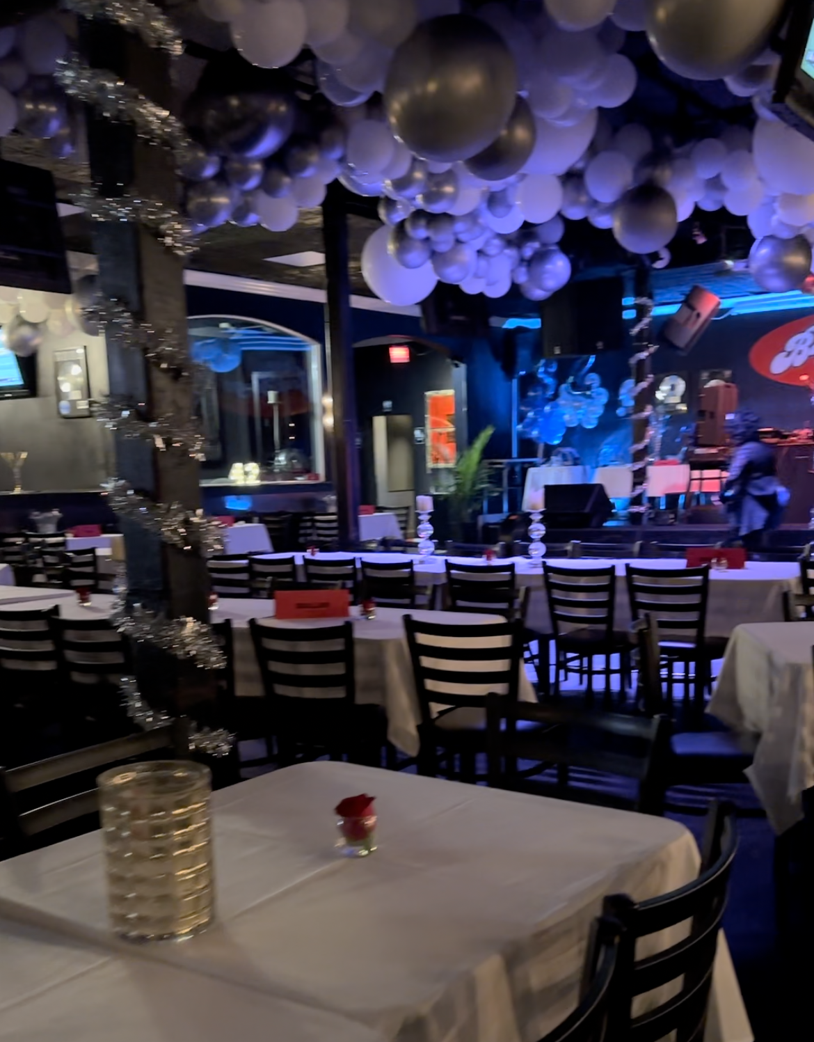 Interior of a restaurant with white tablecloths, black chairs, and a stage in the back. 