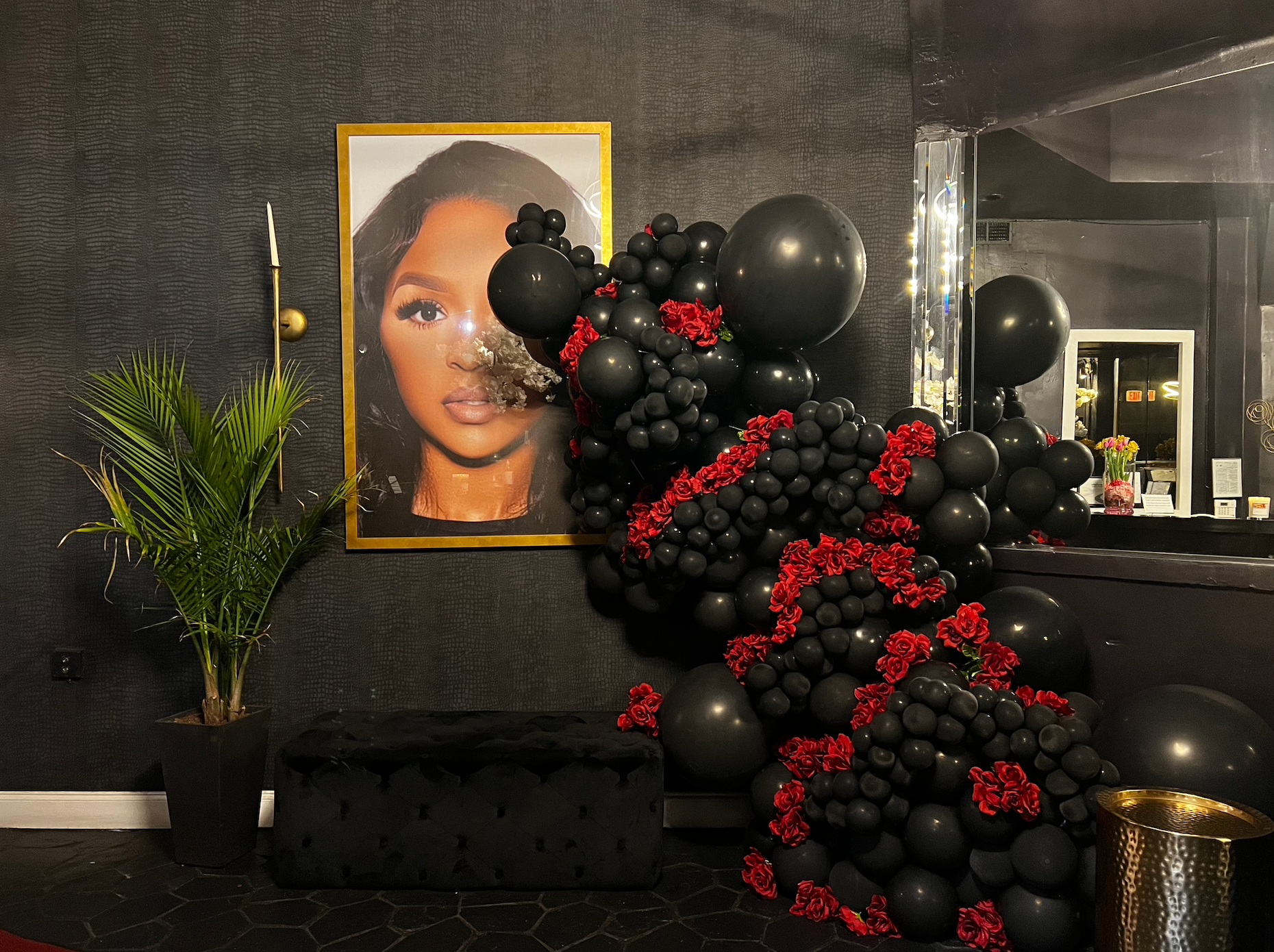 Black balloon garland with red accents next to a portrait, over a black bench and gold table.