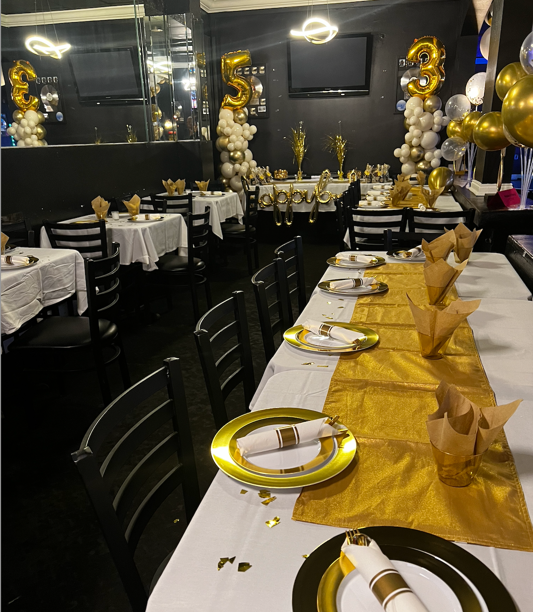 A banquet hall decorated for a birthday celebration with gold and white balloons, tables with gold accents, and empty chairs.