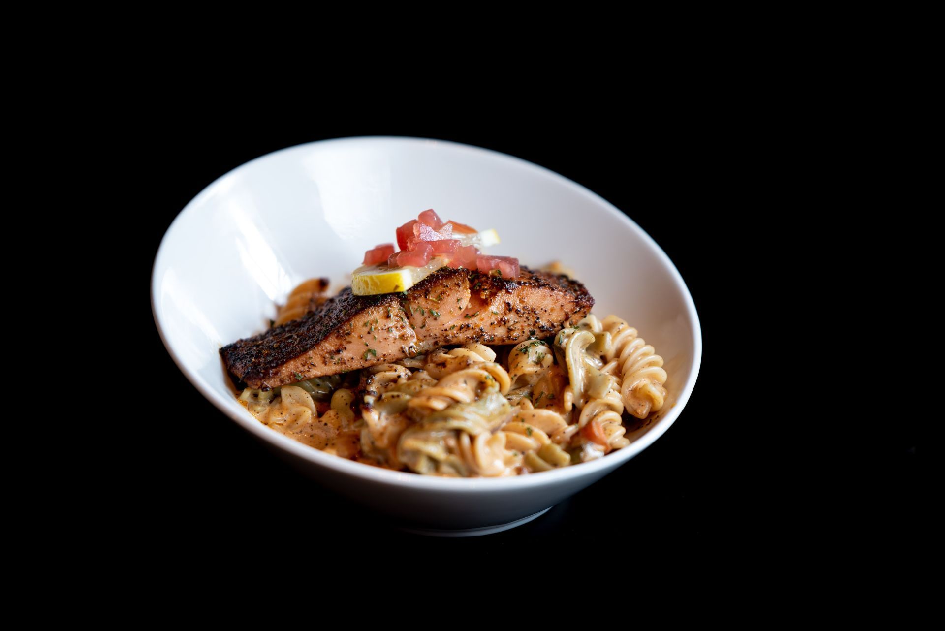 Grilled salmon served over pasta in a white bowl, garnished with tomatoes and lemon. Black background.
