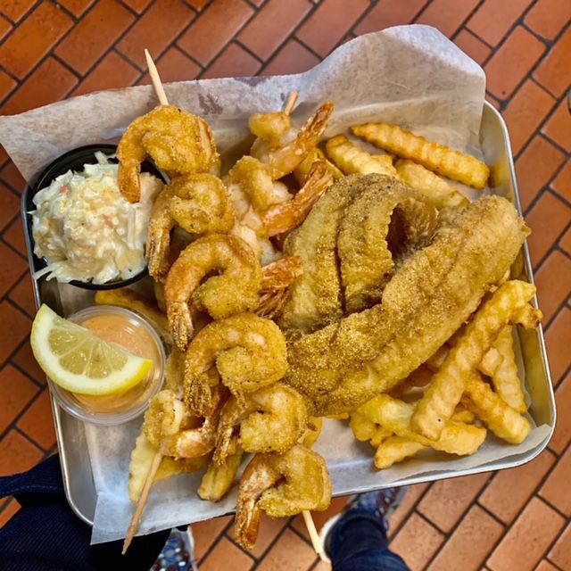 Fried seafood platter with shrimp skewers, fish fillet, fries, coleslaw, lemon, and dipping sauce.