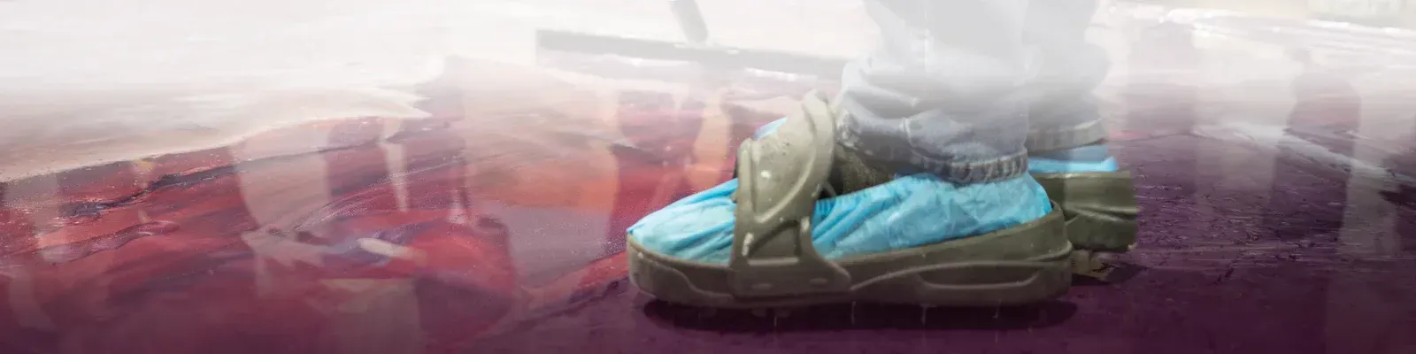 Person's legs and feet wearing blue protective shoe covers and gray clogs on a red surface.