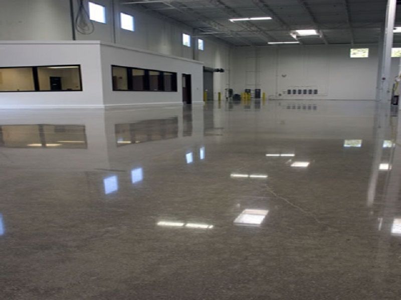 Polished concrete floor in an empty warehouse, reflecting natural light from windows and a small office.