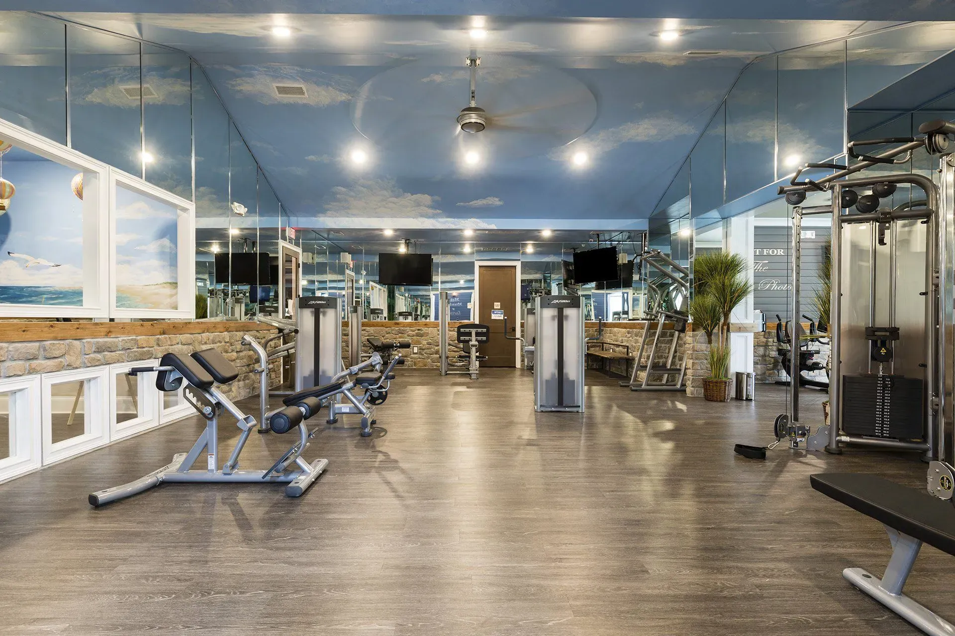 Indoor residential gym with weight machines, cardio equipment, mirrors, and wood-like flooring.