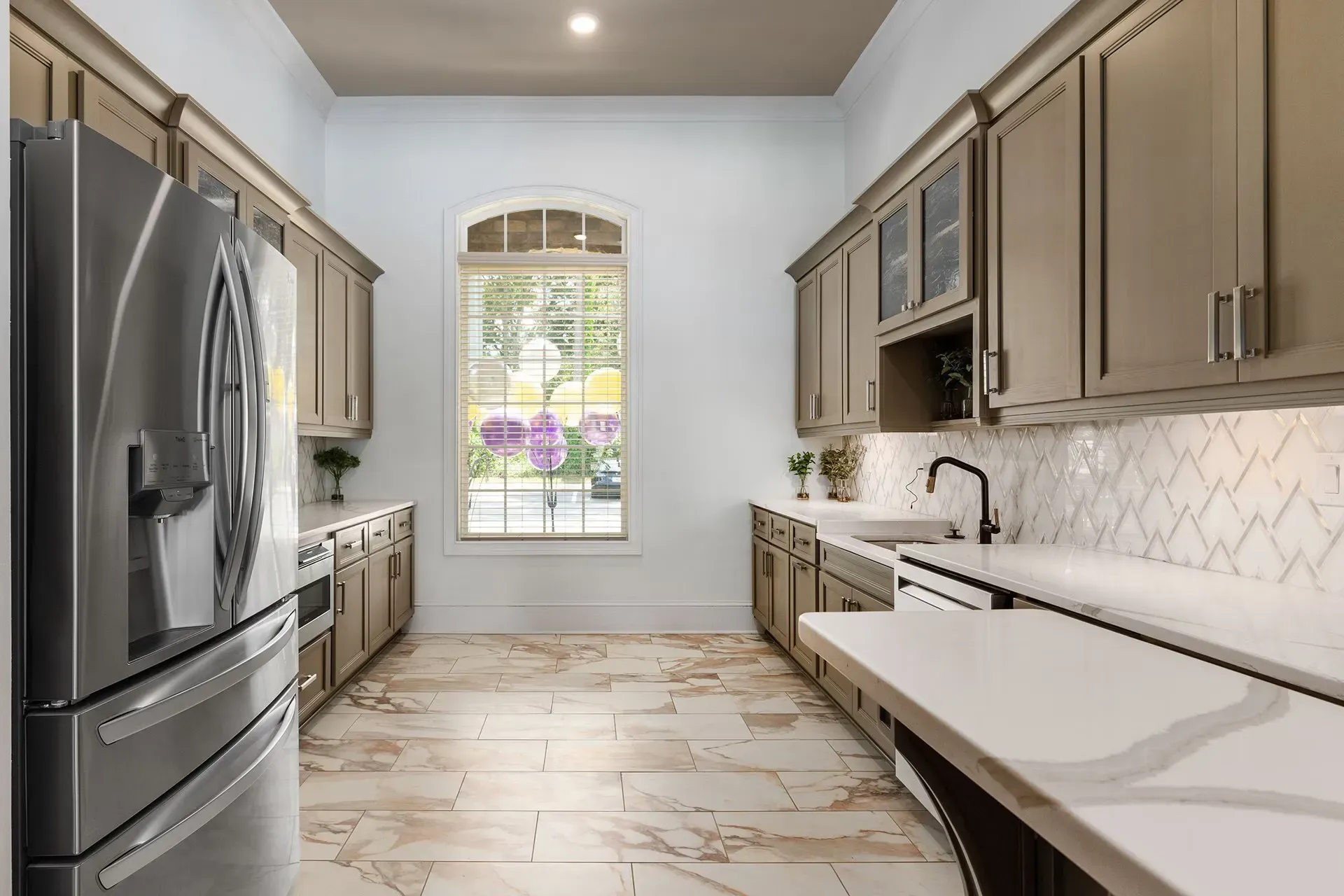 Modern kitchen with stainless steel appliances, beige cabinetry, and marble-like countertops beneath a large window.
