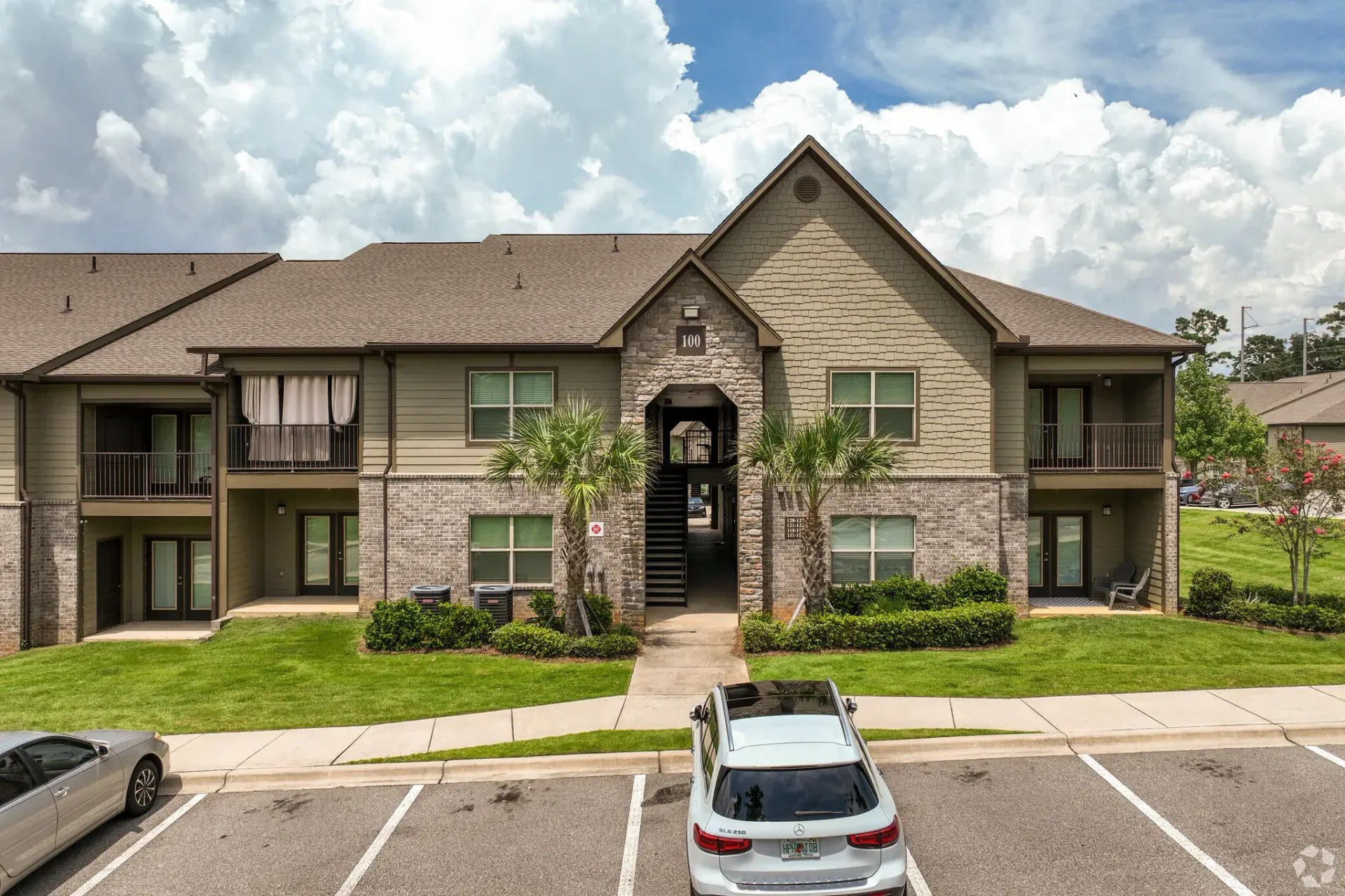 Exterior view of a two-story apartment building with a stone facade, central archway, and palm trees.