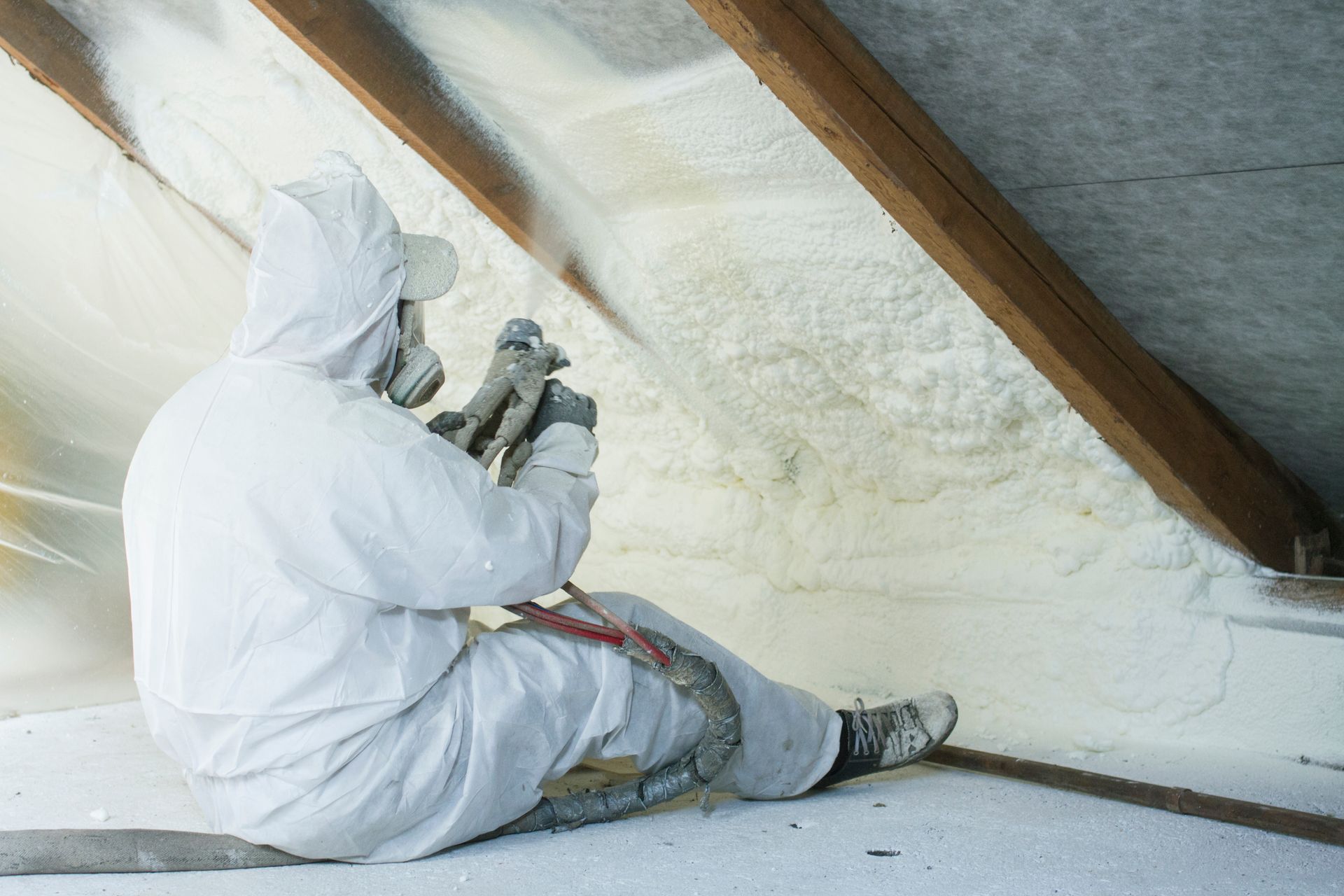 A man in a white suit is spraying insulation in an attic.