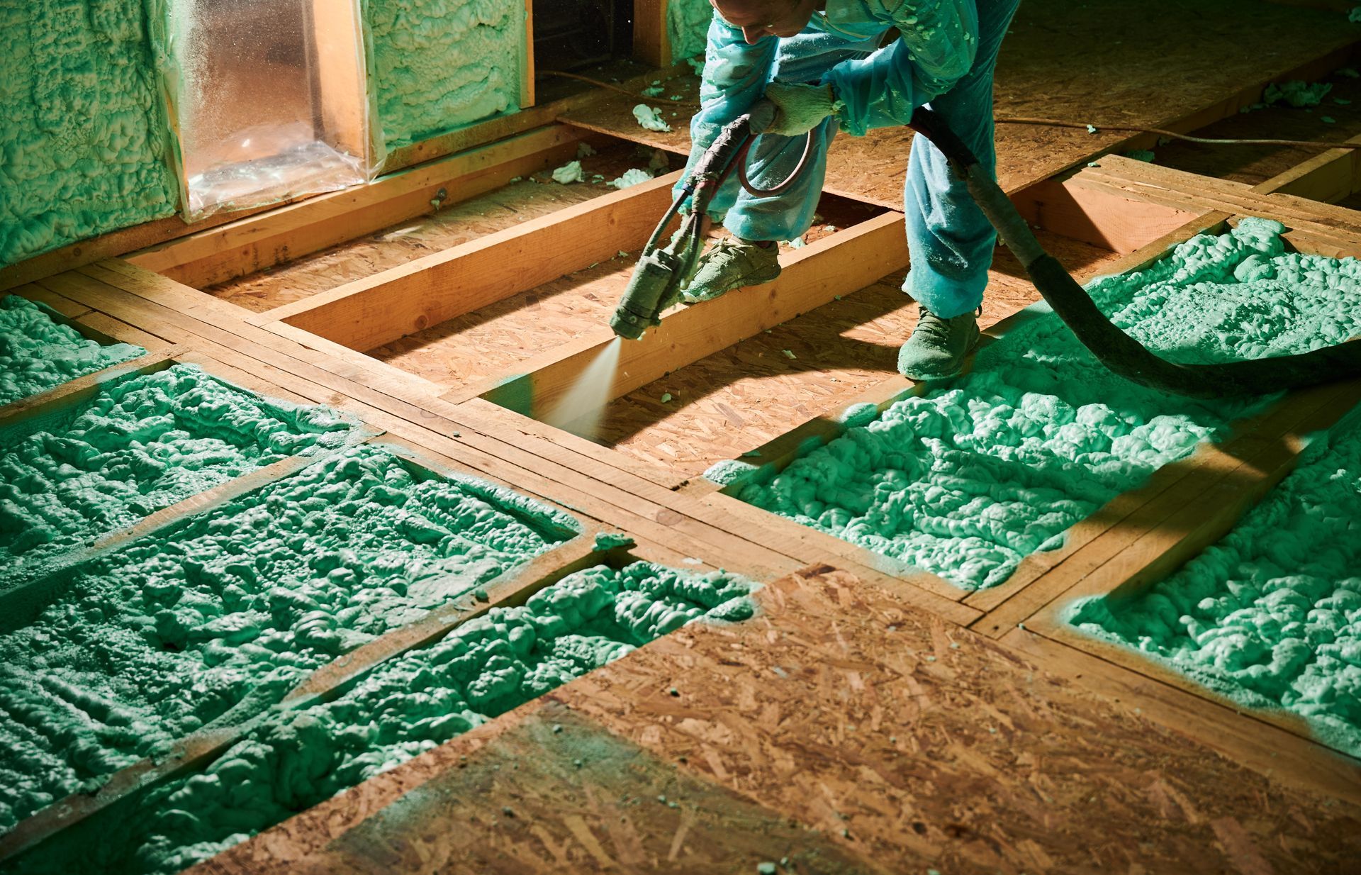 Worker spraying green insulation foam into floor joists in an unfinished attic