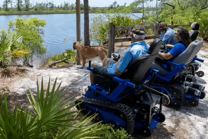 People in blue tracked wheelchairs by a lake, dog, sunny day.