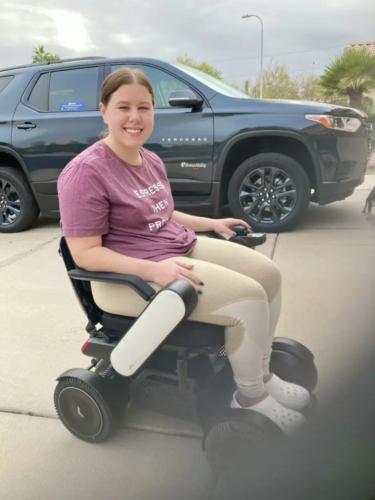 Woman smiling in a powered wheelchair outside a dark SUV. She wears a maroon shirt and khaki pants.