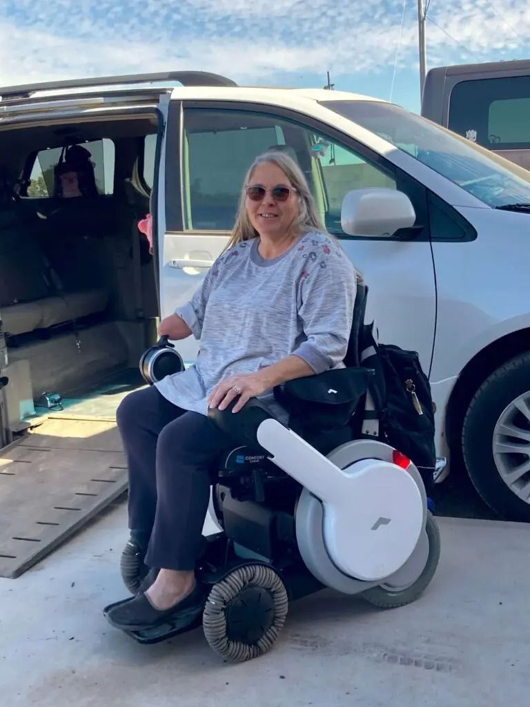 Woman in a wheelchair exiting a vehicle with a ramp. Outdoors, sunny day.