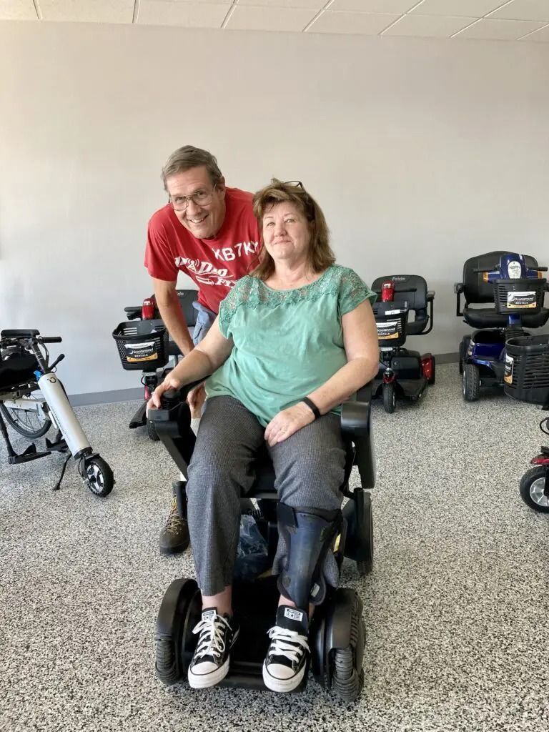 A woman in a wheelchair smiles with a man standing behind her in a mobility scooter showroom.