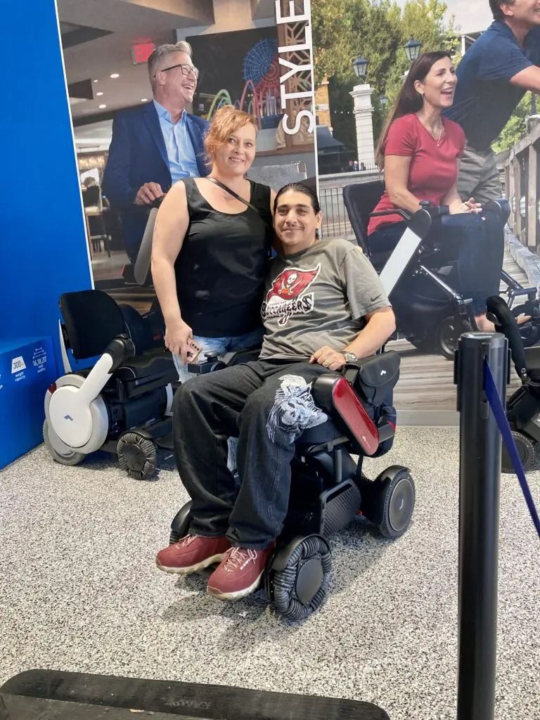 Woman stands beside a man in a power wheelchair, smiling at a trade show with advertising in the background.