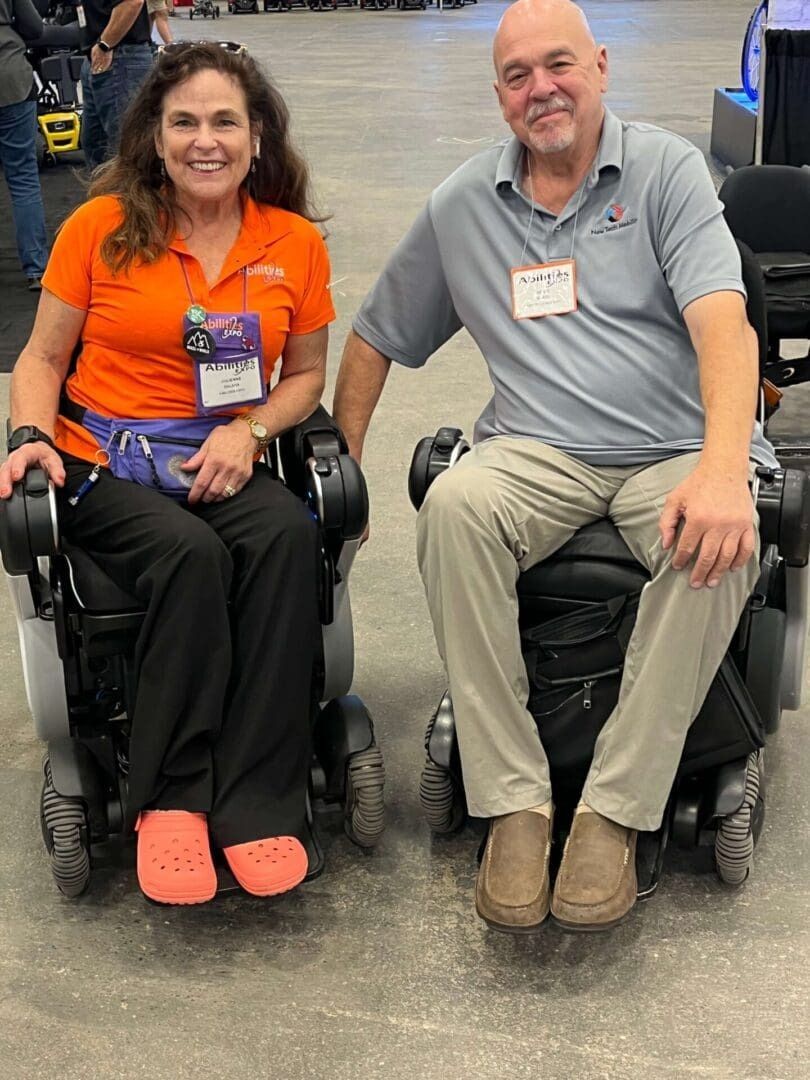 Two people in wheelchairs smile at an event. The woman wears an orange top and Crocs. Man in grey polo, tan pants.