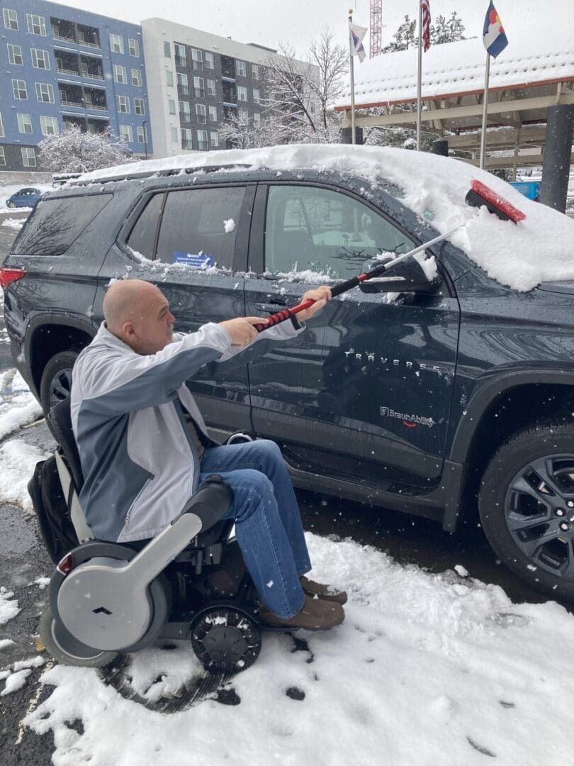Man in wheelchair clearing snow from car windshield with snow brush outside.