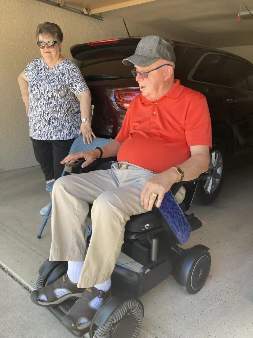 Man in orange shirt on a wheelchair, woman in patterned shirt nearby, both indoors, car in the background.