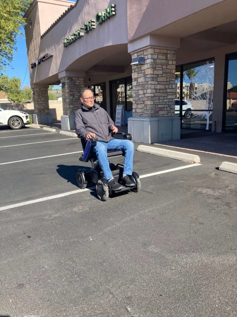 Man in power wheelchair in a parking lot, near a building with glass windows. Sunny day.