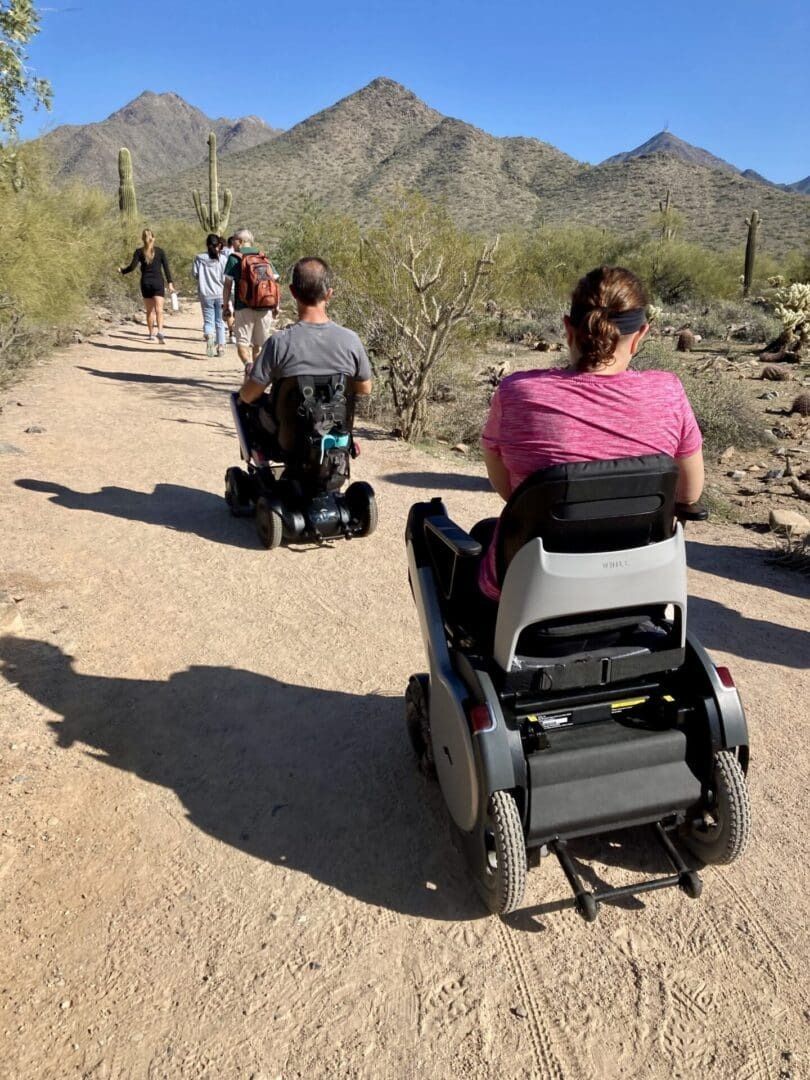 People in wheelchairs and walkers on a desert trail with mountains in the background on a sunny day.