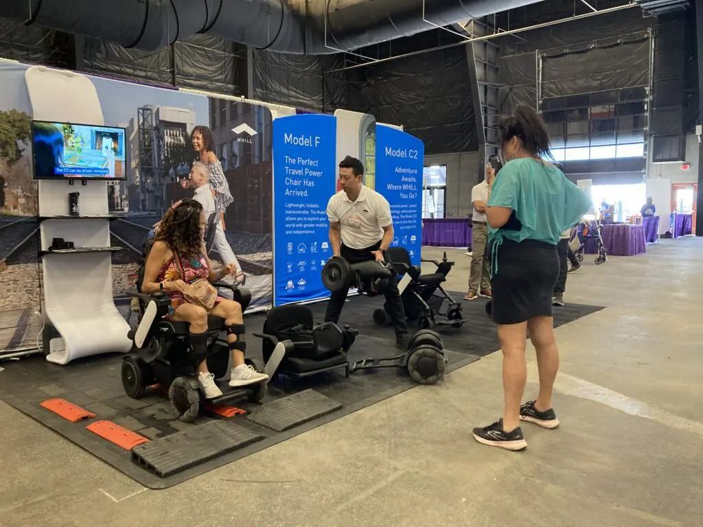 People at an adaptive exercise expo. A woman in a wheelchair, a man holding weights, and another woman watching.