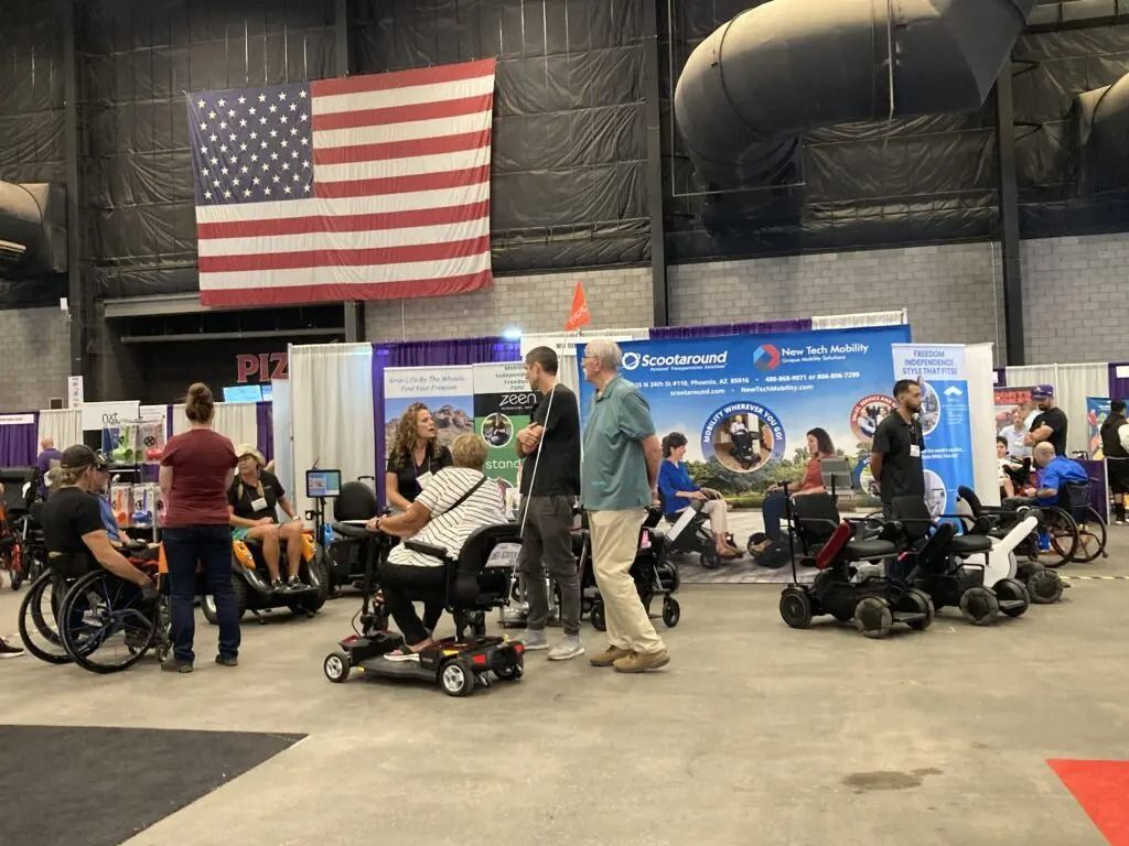 People in wheelchairs and mobility scooters at a convention, with booths, American flag.