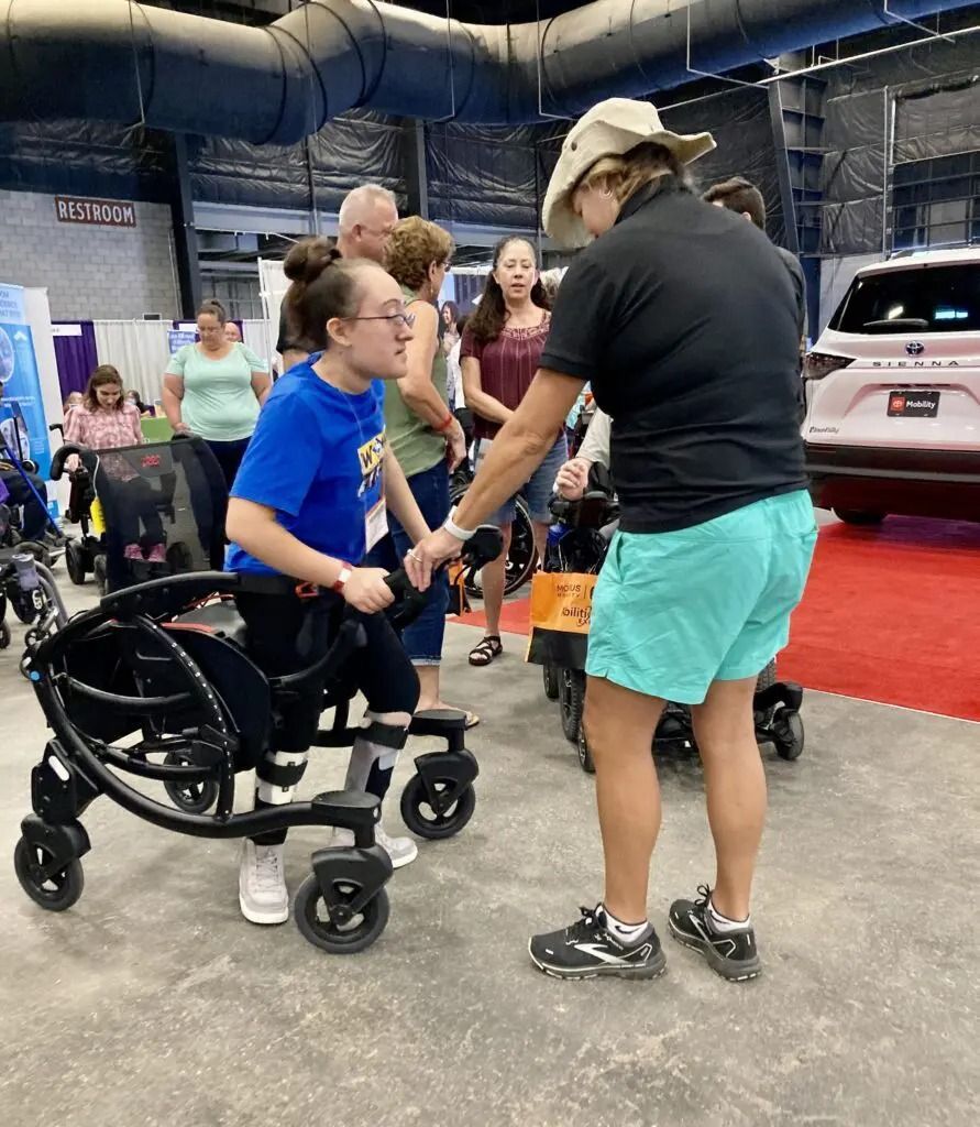 Person in a wheelchair and attendant at an event. Wheelchair is black and foldable. Indoors with other attendees.