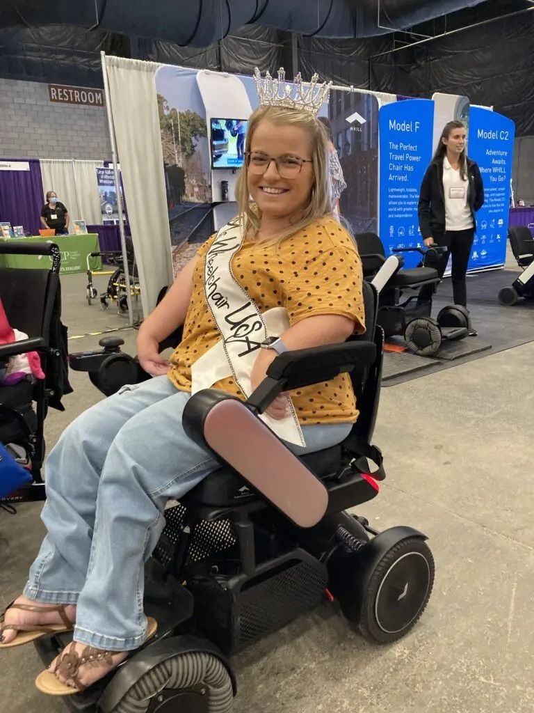 Woman in a wheelchair wearing a crown and sash smiles at an event.  Behind her are vendor booths.