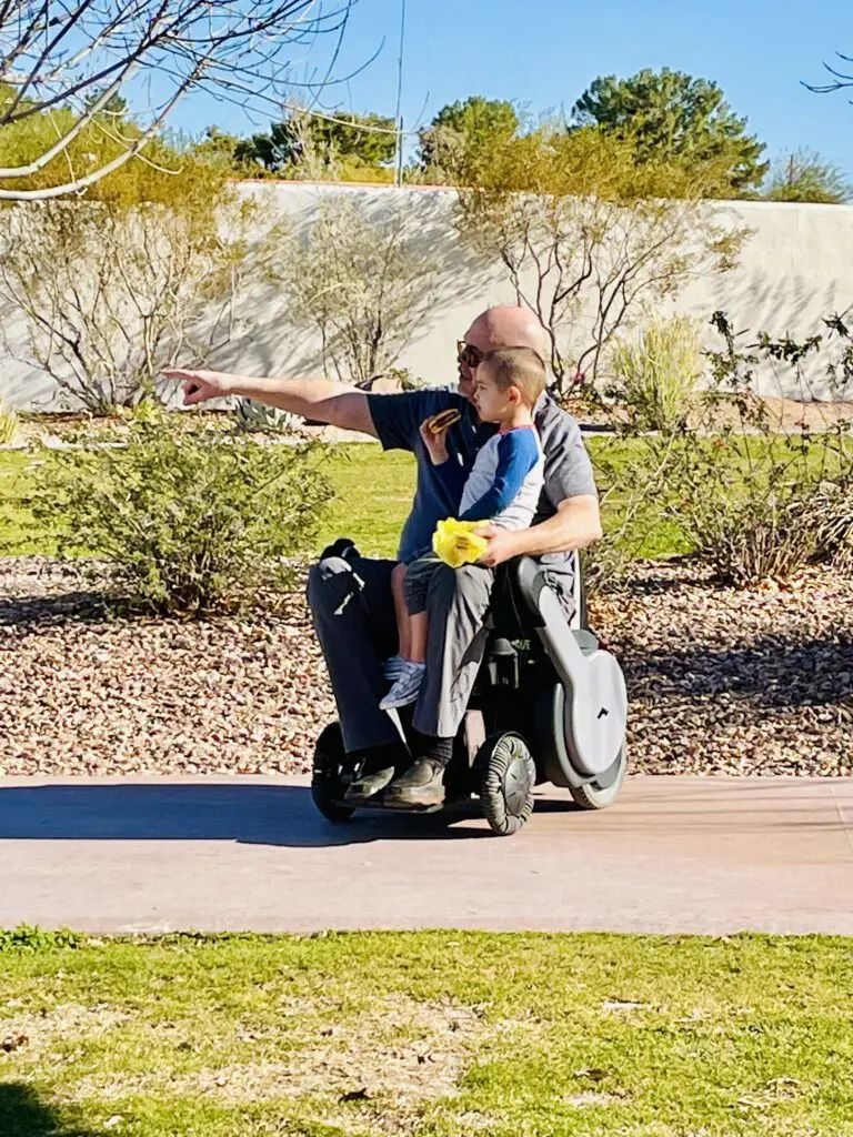 Man in wheelchair points, holding a child outdoors. Green grass, sunny day, white wall, trees in background.