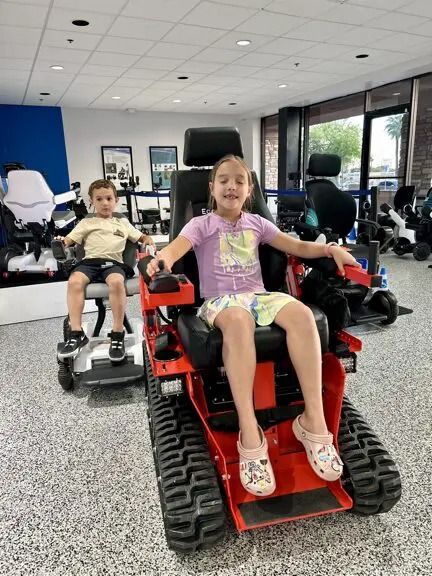 Two children in wheelchairs, smiling. One in red, heavy-duty chair, another in a white chair. Indoor setting.