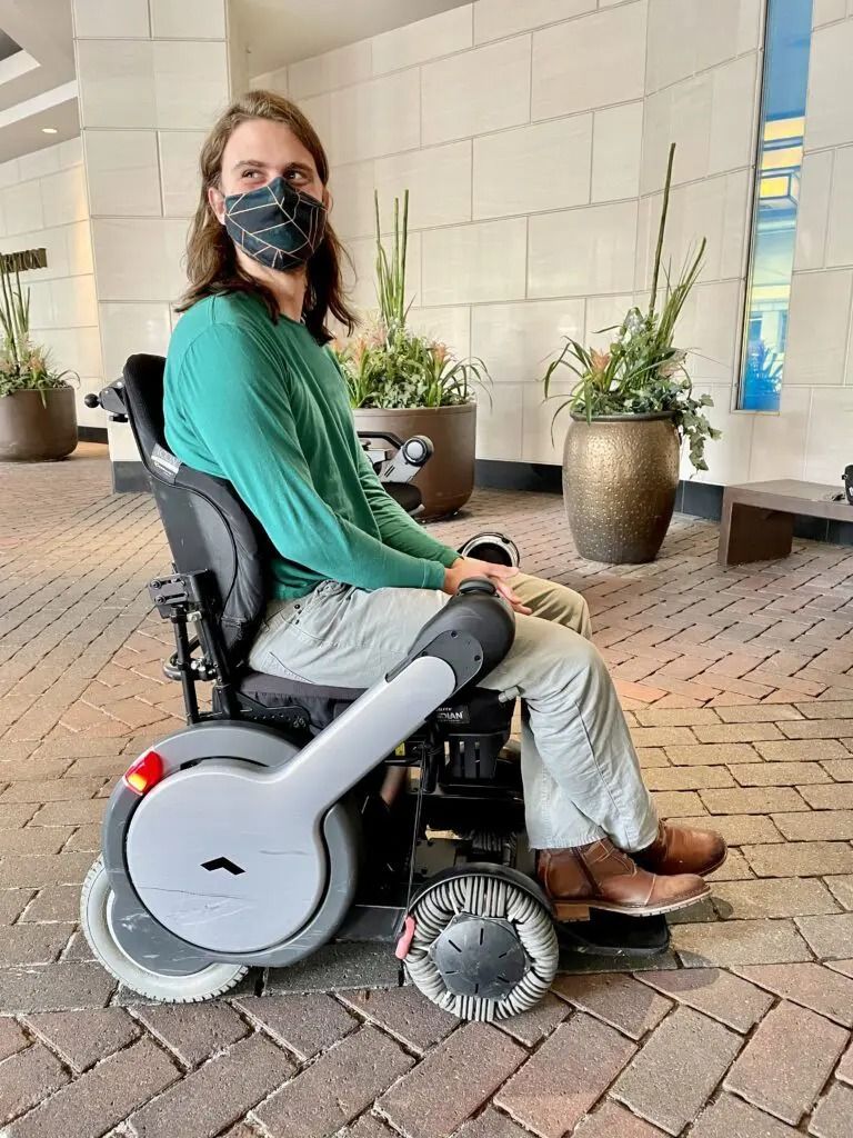 Person in wheelchair wearing a mask, outdoors. Light-skinned with long brown hair, wearing green shirt and khaki pants.