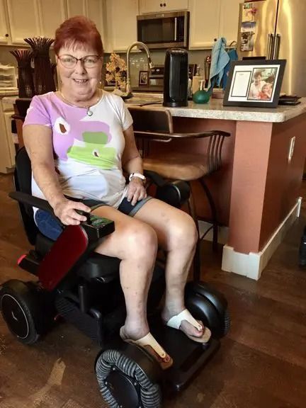 Woman in a wheelchair smiling, in a kitchen. She wears glasses, a colorful shirt, and shorts.