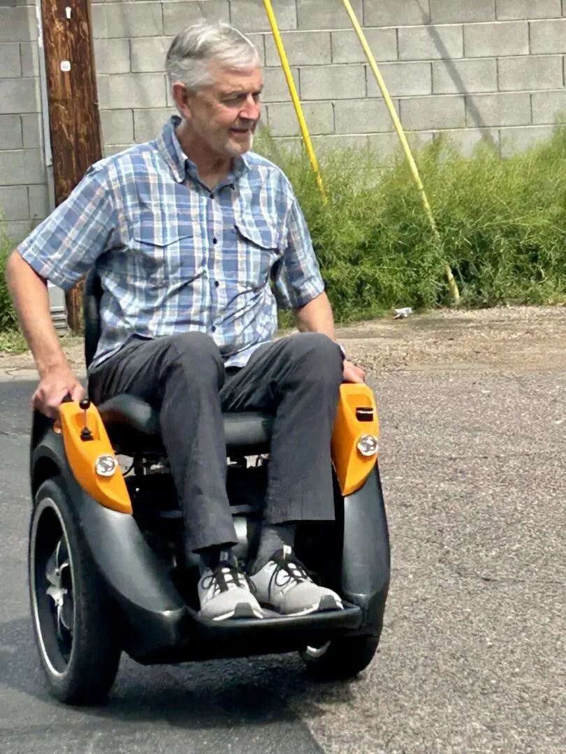 Man in plaid shirt on black and orange power wheelchair on a paved road, next to a building.