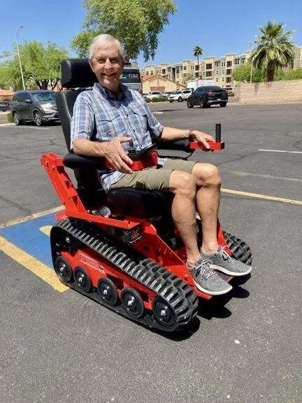 Man in a red track chair smiles outdoors in a parking space, blue sky.
